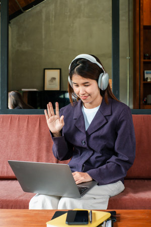 Woman wearing headphones, waving at laptop during a video call, seated on a sofa in a contemporary living room.の写真素材