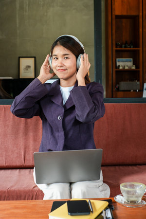 A woman wearing headphones, sitting with a laptop in a modern cafe, enjoying music and working.の写真素材