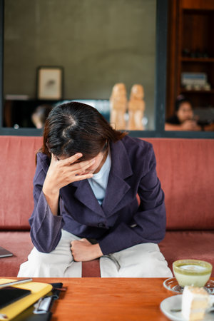 Woman in a cafe looking stressed, sitting with hand on forehead, reflecting in a cozy setting.の写真素材