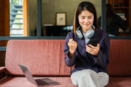 A young woman with headphones celebrates success while using her smartphone and laptop in a modern home office.の写真素材