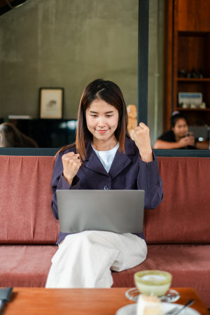 A joyful young woman celebrates a success while working on her laptop in a cozy cafe environment.の写真素材