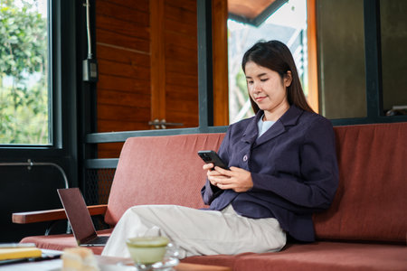 A young woman sits comfortably on a sofa, using her smartphone in a modern living room with wooden decor and natural lighting.の写真素材