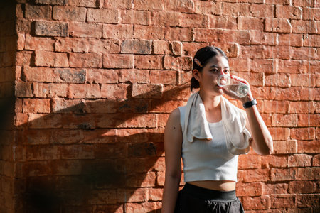 Athletic woman in sportswear hydrating post-exercise, leaning on a sunlit brick wall.の写真素材