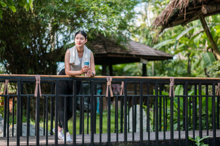 A woman enjoys a refreshing drink while leaning on a railing in a lush tropical garden, surrounded by greenery and bamboo structures.の写真素材