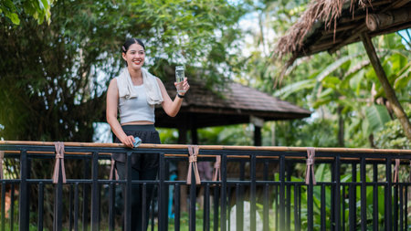Smiling woman holding a glass of water on a balcony, set against a backdrop of tropical plants and wooden structures.の写真素材