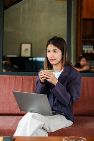 A woman in a cafe holding a cup, working on her laptop, creating a relaxed and productive atmosphere.の写真素材