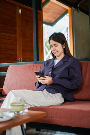 A young woman sits comfortably on a sofa, focused on her smartphone in a stylish cafe with a wooden interior.の写真素材