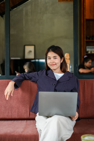Woman sitting on a sofa with a laptop in a modern cafe, exuding confidence and relaxation.の写真素材