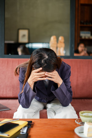 A woman in a blazer sits on a couch with her head in her hands, conveying stress or contemplation in a modern cafe environment.の写真素材