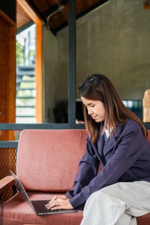 A young woman in a blazer works on her laptop in a stylish, modern home setting with natural light and comfortable furniture.の写真素材