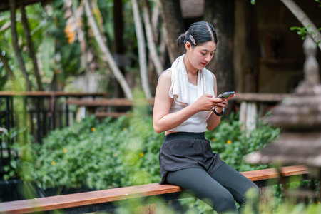 A young woman sits on a wooden railing in a garden, focused on her smartphone, surrounded by lush greenery.の写真素材