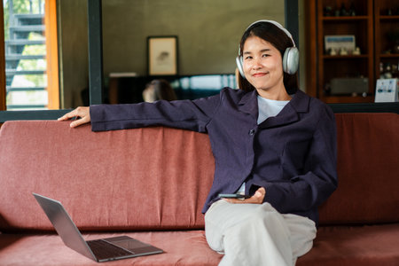 A woman wearing headphones sits on a sofa, enjoying music. A laptop and smartphone are nearby, creating a relaxed and modern atmosphere.の写真素材
