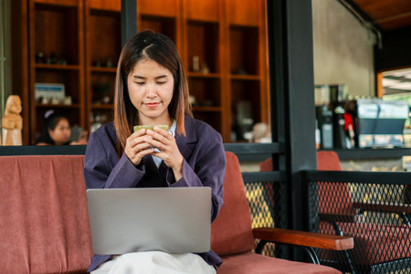 A woman in a cafe enjoying coffee while working on her laptop, surrounded by a cozy and modern wooden interior.の写真素材