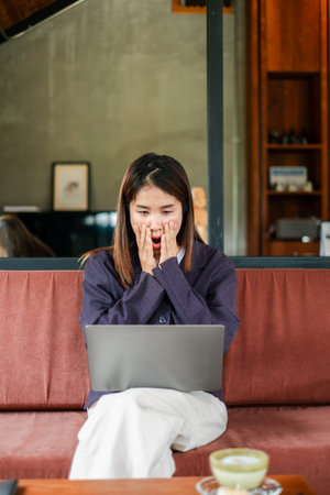 A woman expresses excitement while using a laptop on a couch in a stylish living room with wooden decor and natural lighting.の写真素材