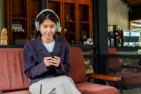 A young woman enjoys music on headphones while using her smartphone in a stylish cafe environment.の写真素材