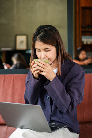 A woman in a cafe savoring a hot drink while using her laptop, creating a warm and productive atmosphere.の写真素材