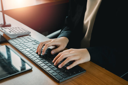 Close-up of hands typing on a keyboard in a modern office environment with sunlight streaming in.の写真素材