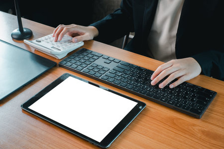 Business professional using a tablet, keyboard, and calculator at a wooden desk in a modern office setting.の写真素材