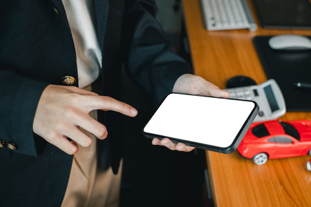 Businessperson pointing at smartphone with blank screen on office desk, featuring calculator and toy car.の写真素材