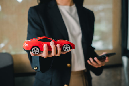 A business professional in a suit holds a red toy car and a smartphone, symbolizing innovation and technology in the automotive industry.の写真素材