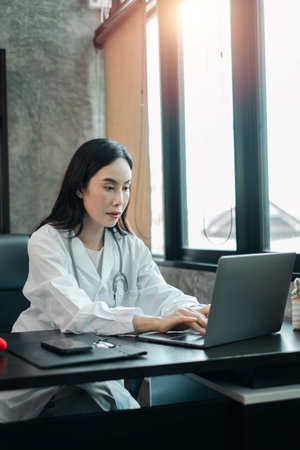 Female doctor in white coat working on a laptop in a modern office setting with natural light.の写真素材