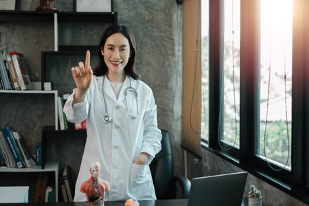Female doctor in a modern office, gesturing number one, with anatomical model and laptop on desk.の写真素材