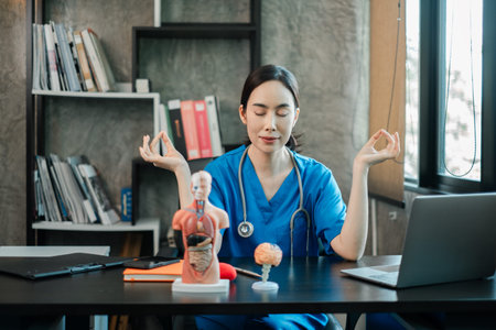 Female doctor in blue scrubs meditating at desk in a modern office, promoting mindfulness and stress...