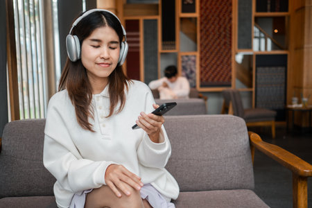 Woman wearing headphones, relaxing in a cafe, enjoying music on her smartphone in a comfortable and modern environment.の写真素材