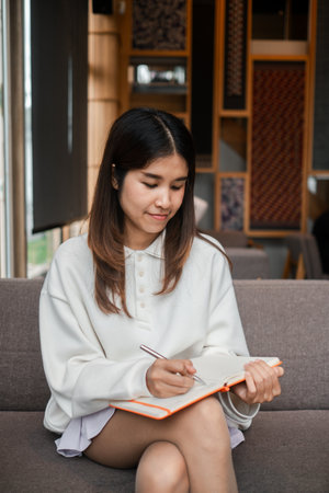 A young woman sits on a sofa, writing in a notebook, surrounded by a modern and cozy interior setting.の写真素材