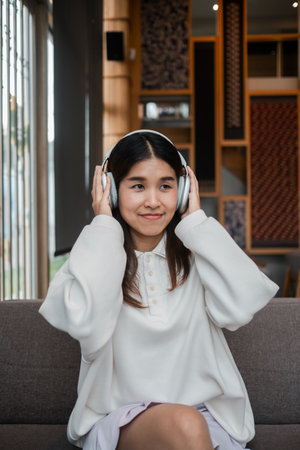 A young woman wearing headphones, sitting on a sofa, enjoying music in a stylish and cozy living room.の写真素材