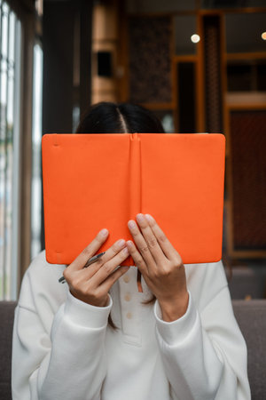 A woman in a white sweater holds an orange notebook in a stylish cafe with natural light and modern...