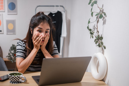 Young woman in striped shirt looking surprised at laptop in a modern home office setting with...