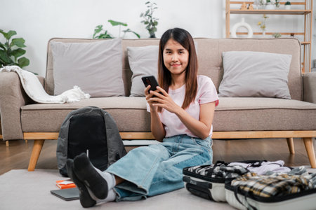 Woman sitting on floor with smartphone, packing suitcase for travel in a cozy living room setting.の写真素材