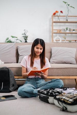 Young woman sitting on floor, writing in notebook, preparing for travel. Modern living room setting with backpack and suitcase.の写真素材