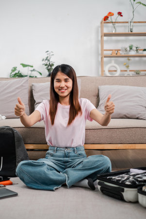 Smiling woman sitting on floor, packing suitcase, giving thumbs up in cozy living room setting.の写真素材