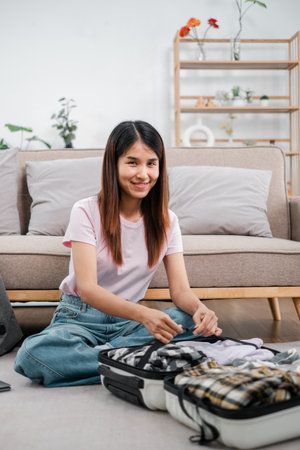 Smiling young woman packing clothes into a suitcase in a cozy living room, preparing for a travel adventure.の写真素材