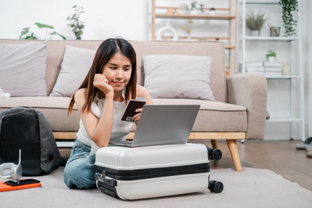 Woman using laptop and credit card for online shopping, sitting on suitcase in a modern living room.の写真素材