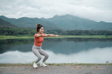 Woman performing squats by a lake with mountains in the background, showcasing outdoor fitness and natural beauty.の写真素材