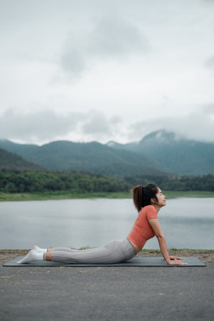 A woman in activewear performs yoga by a tranquil lake, surrounded by lush mountains under a cloudy...