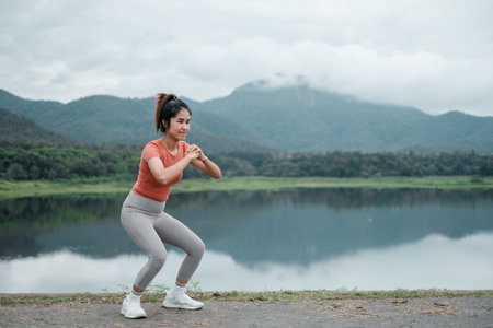 Woman performing squats by a lake with mountains in the background, showcasing outdoor fitness and nature.の写真素材