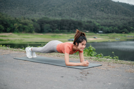 Woman in athletic wear doing a plank exercise on a yoga mat by a lake with green hills.の写真素材