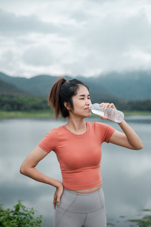 A young woman in athletic wear drinks water by a lake, surrounded by mountains and nature.の写真素材
