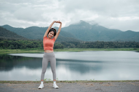 Woman in activewear stretching by a lake with mountains in the background, showcasing fitness and...