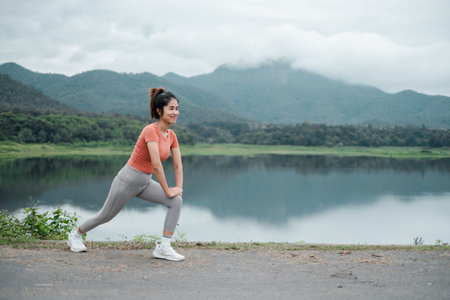 Woman in activewear stretching by a lake with mountains in the background, showcasing fitness and...