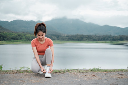 Woman in activewear tying shoelaces by a lake with mountains in the background, embracing nature and fitness.の写真素材