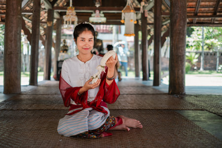 A woman in traditional Thai clothing sits in a wooden pavilion, holding a floral garland, showcasing...