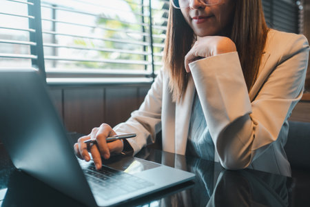 Businesswoman in a white blazer using a laptop in a contemporary office setting with natural light.の写真素材