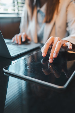 Businesswoman multitasking with tablet and laptop in a modern office environment, showcasing digital communication and productivity.の写真素材