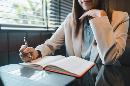 Professional woman taking notes in a notebook at a modern office desk with a laptop, showcasing productivity and focus.の写真素材