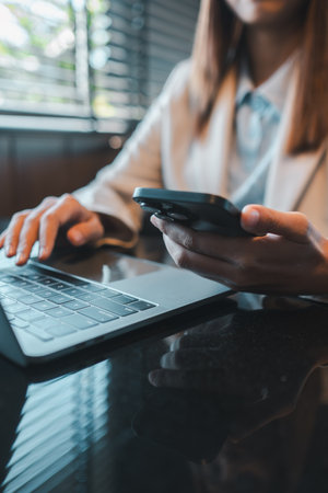 Professional woman multitasking with smartphone and laptop at a sleek office desk, emphasizing modern work dynamics.の写真素材